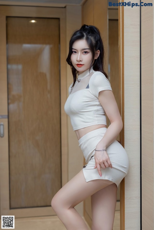 A woman in a white top and skirt leaning against a wall.