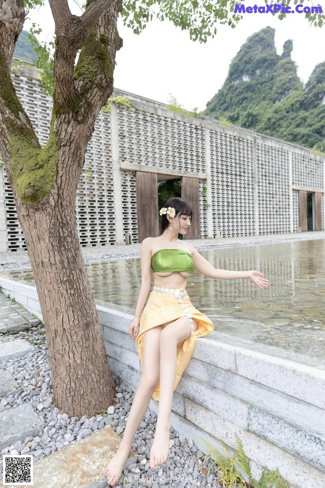 A woman in a green top and yellow skirt sitting by a tree.