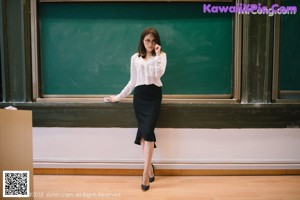 A woman sitting on top of a desk next to a blackboard.